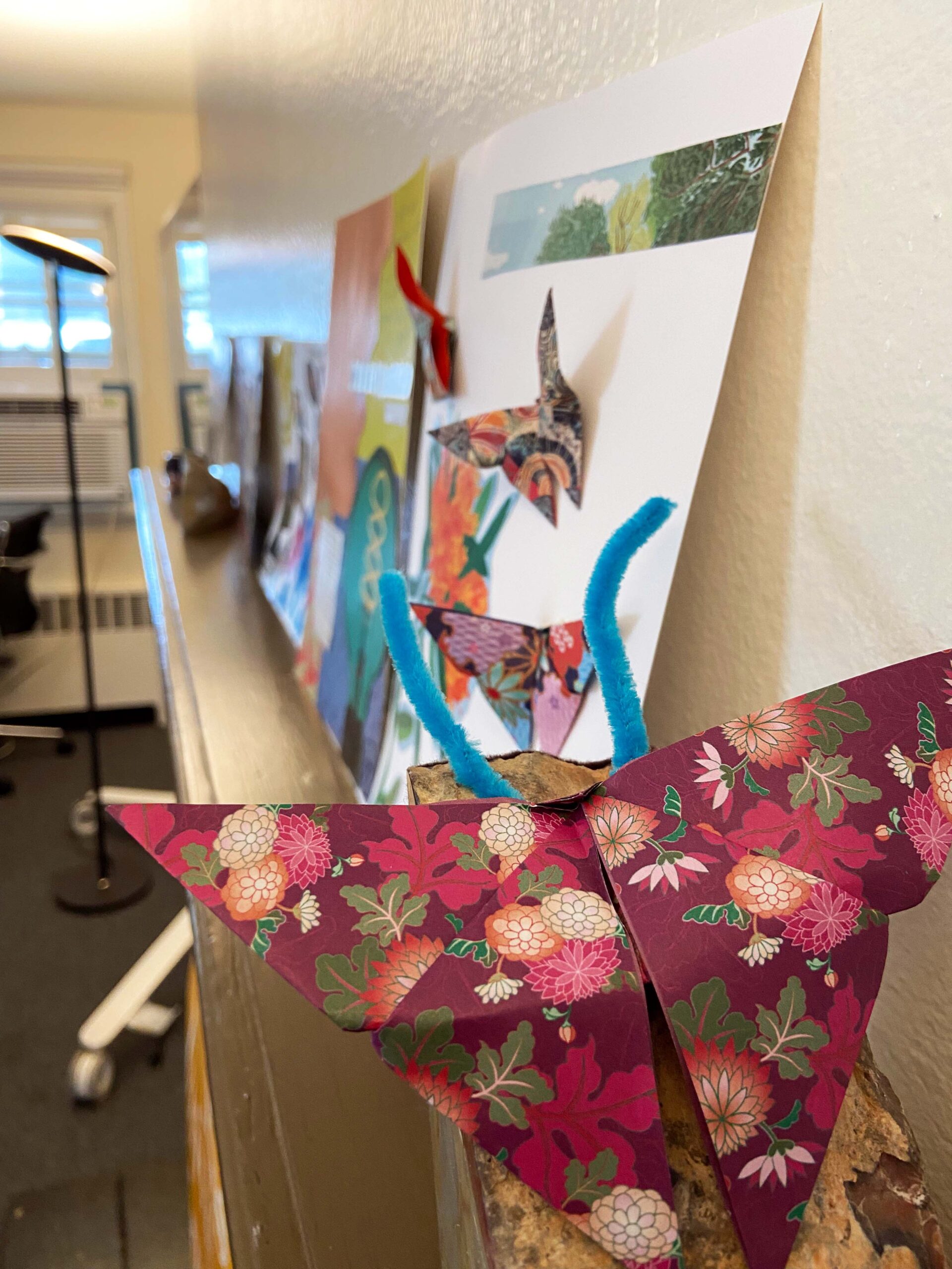 1. An origami butterfly made from maroon wrapping paper (featuring leaves and chrysanthemums), with blue antennae, sits on a piece of wood. Ahead of the butterfly is a ledge on which a couple of images are placed. One can see more butterflies on the surface of the images.