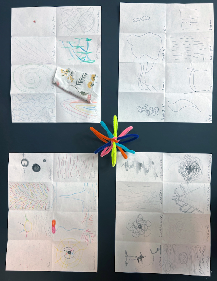 four sheets of paper rest on a black table top. A colorful pinwheel made of pipe cleaners rests in the middle. Each of the papers has pencil drawings featuring various organic shapes and objects.