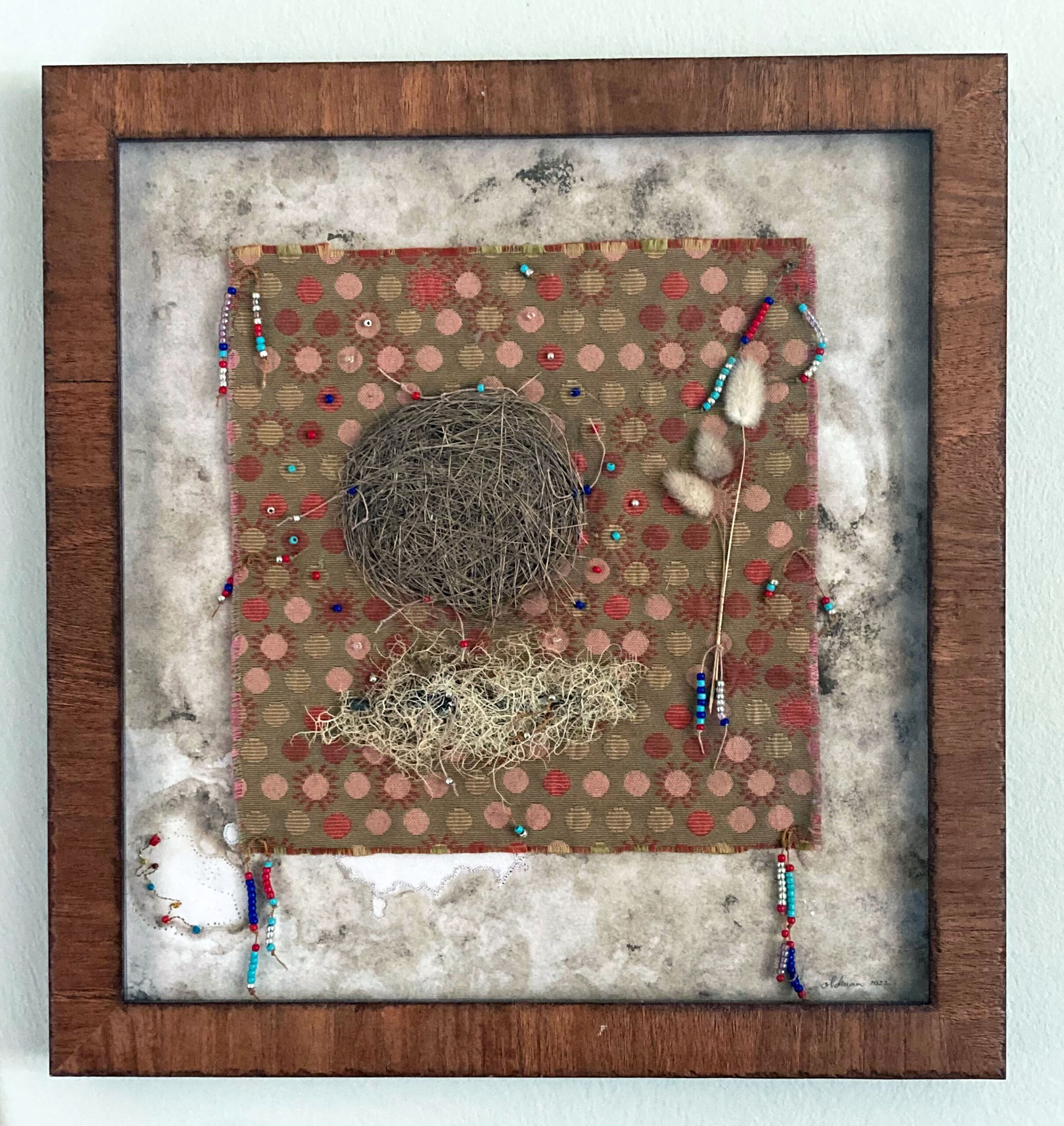 A square wooden frame. In the center of the frame is another square. A bronze piece of fabric with red, pink, cream, and brown, circles and suns. There are beads strung through parts of the fabric. The soft tufts of stalks of grass rests to the right. To the left, are a couple of nests. Around the fabric square is a band of white that is torn and colored by soil.