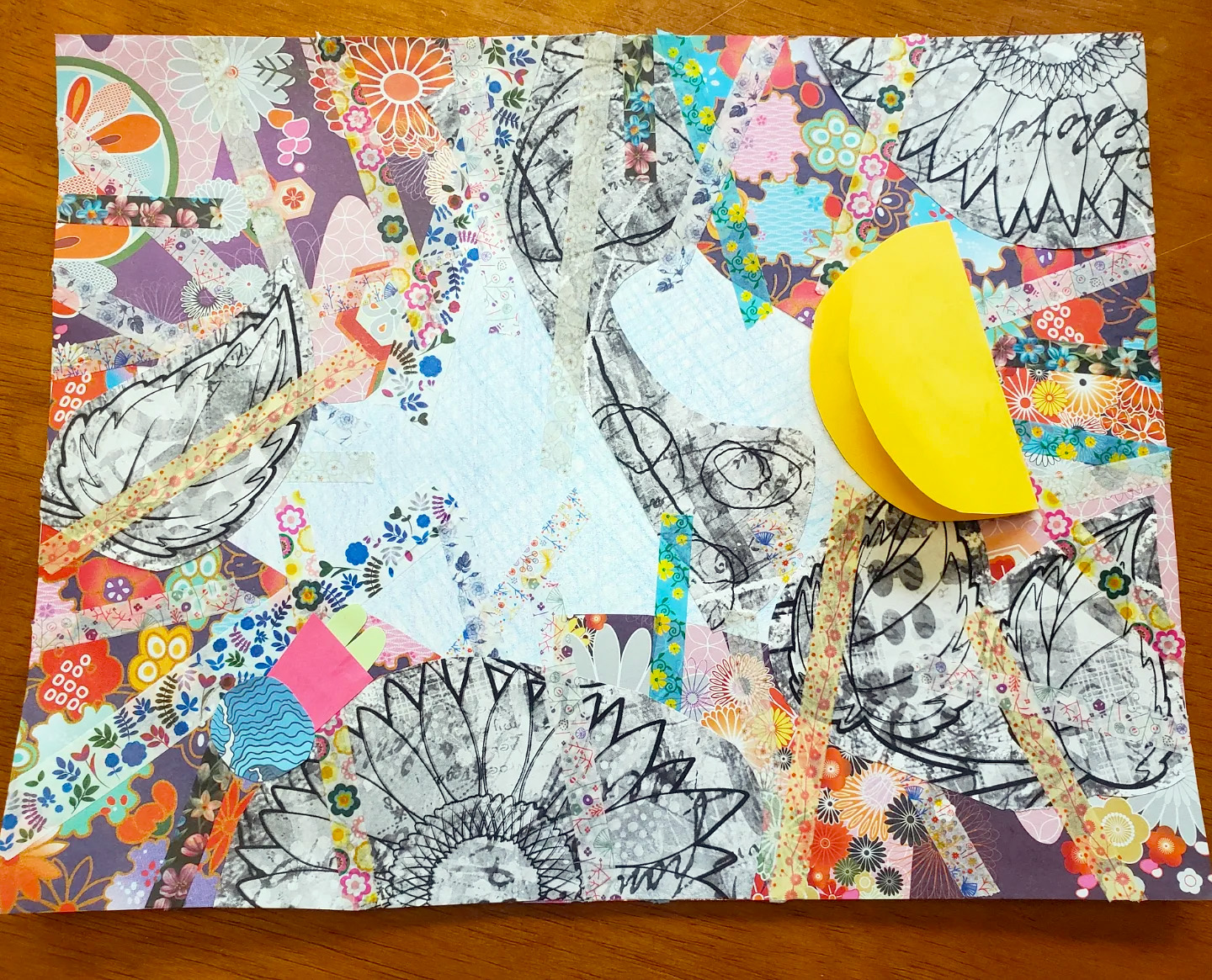 A collage rests against a wooden table, which can be seen along the edges of the image. The image itself is a collage of decorative floral wallpaper, with cutouts of line drawings of leaves and flowers. There are strips of floral paper across the image as well. A folded bright yellow moon rests toward the top right of the image.