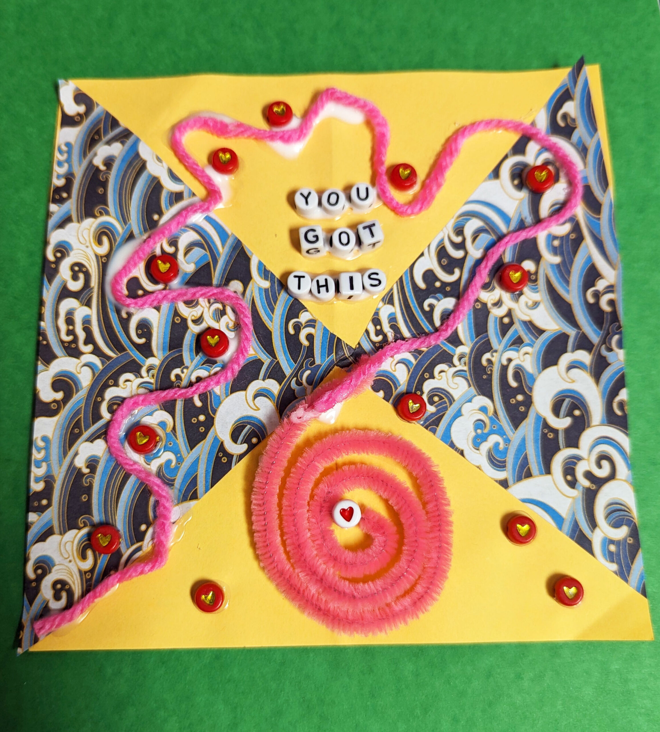 Against a green background is a collage. In the middle is a square composed of four triangles: two yellow on the top and bottom, and two featuring Japanese style waves on the left and right. A pink string weaves over the image. It is surrounded by red beads which have a golden heart at the center. The string leads to a circle of pink (made with a pipecleaner) which has a white bead at its center with a red heart on it. On the top quadrant are the following words, made with beads: You got this.